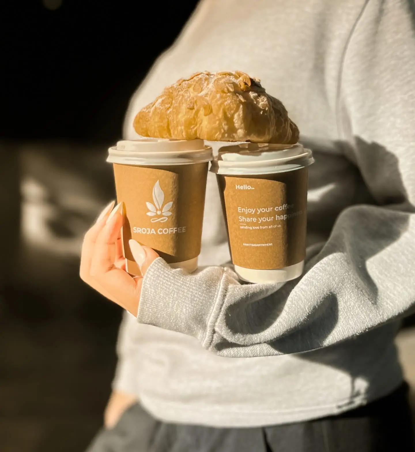 Hands holding two branded coffee cups and a bun, café feed content